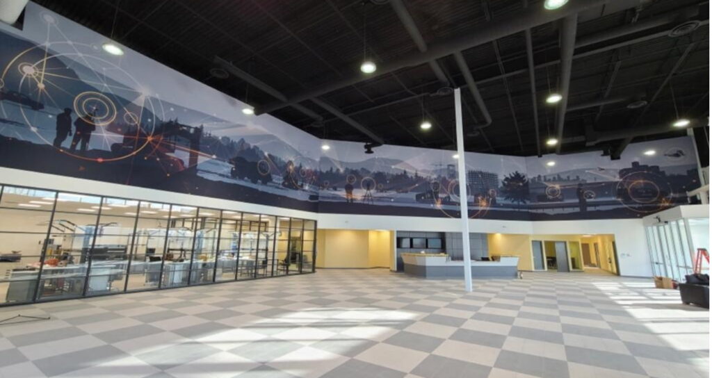 A modern, spacious hall features a panoramic mural depicting industrial and technological scenes with abstract overlays, a glass-walled lab, and a checkered floor.