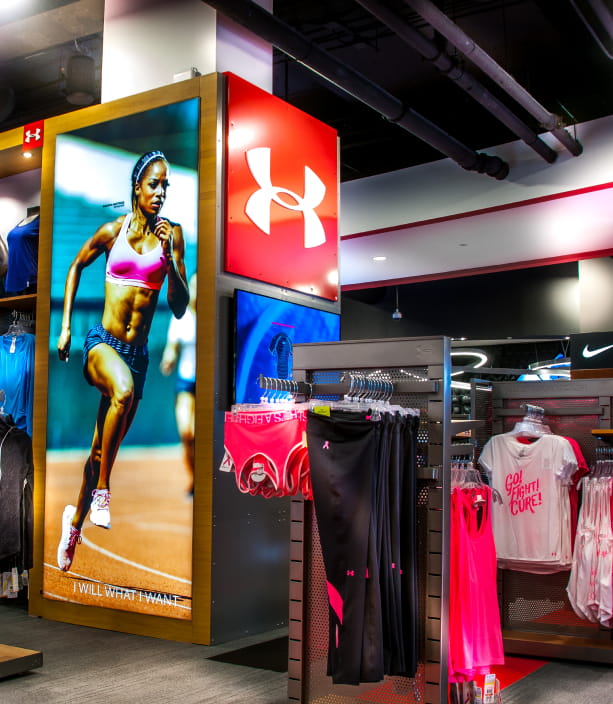 "An Under Armour retail store interior with a large illuminated sign featuring the UA logo, a female runner on a display, and racks of athletic apparel."