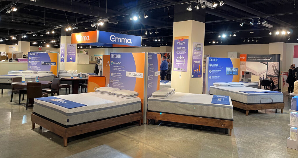 A bright, spacious mattress showroom displays several beds on wooden frames, accompanied by large promotional signs and brand banners.