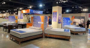 A bright, spacious mattress showroom displays several beds on wooden frames, accompanied by large promotional signs and brand banners.