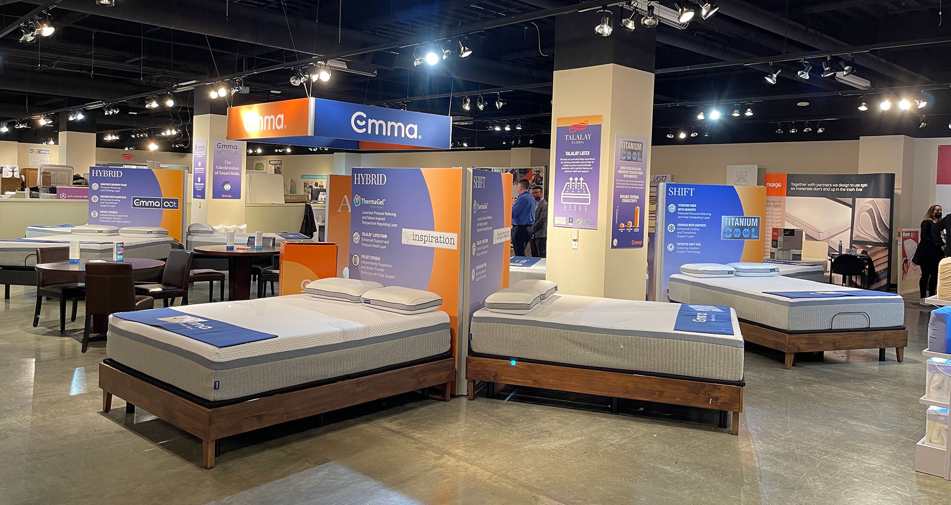 A bright, spacious mattress showroom displays several beds on wooden frames, accompanied by large promotional signs and brand banners.