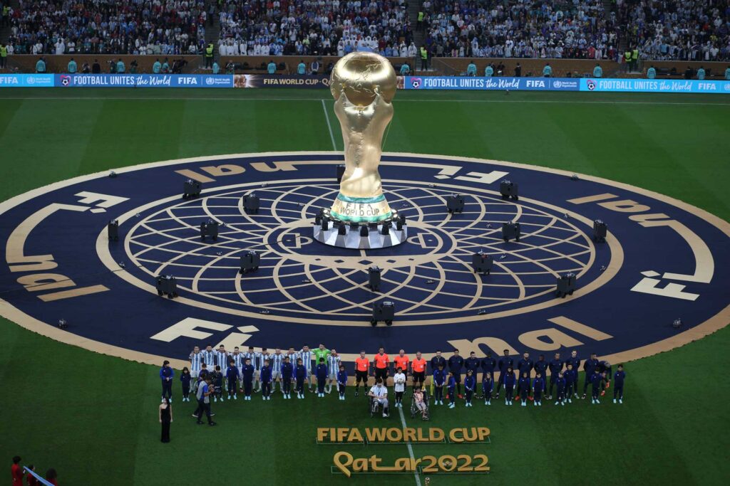 The golden FIFA World Cup Qatar 2022 trophy stands centrally on a decorated pitch, flanked by two football teams and overlooked by a stadium full of spectators.
