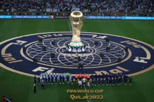The golden FIFA World Cup Qatar 2022 trophy stands centrally on a decorated pitch, flanked by two football teams and overlooked by a stadium full of spectators.