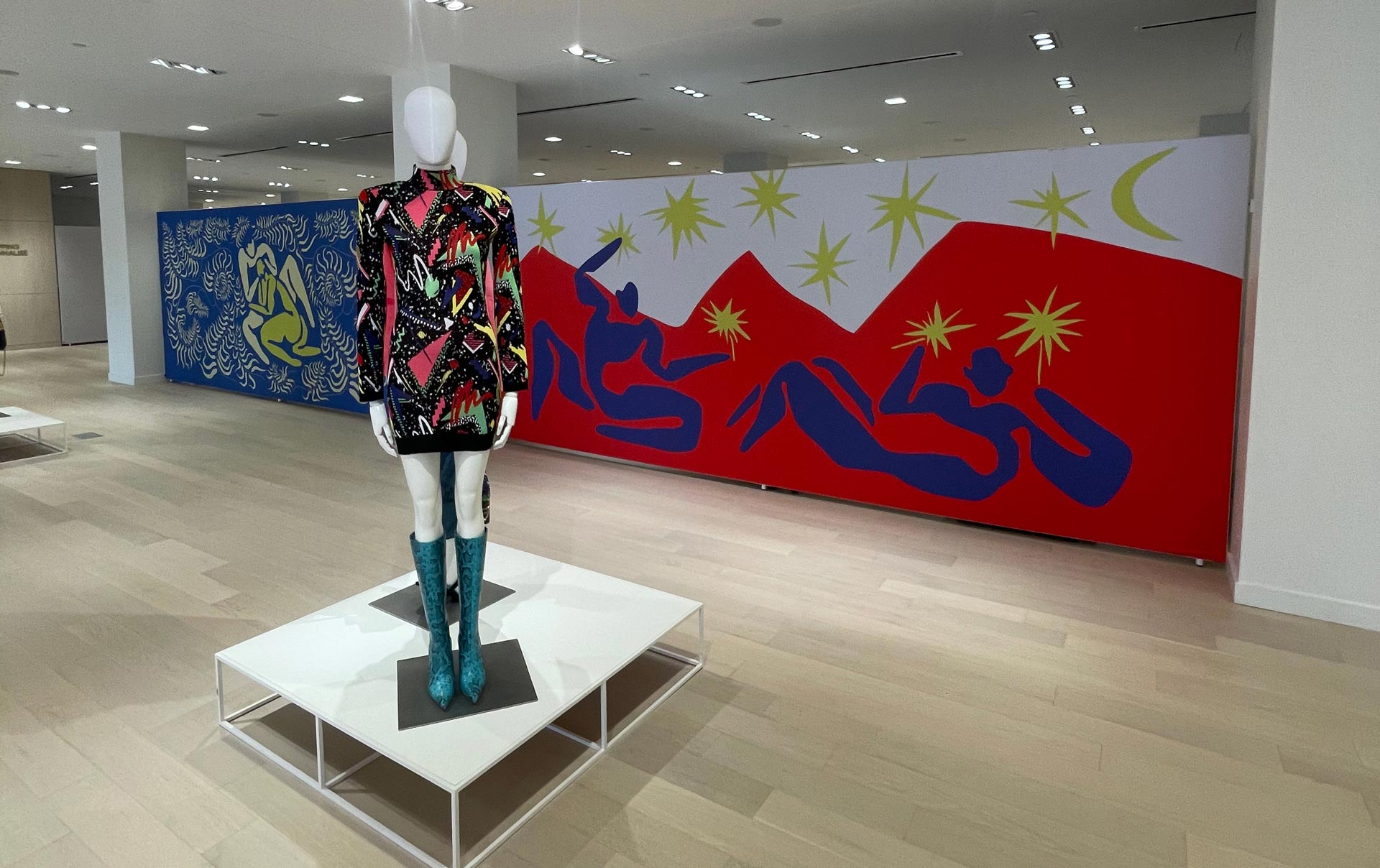 A mannequin in a vibrant patterned dress and teal boots stands before a wall covered in two large, colorful Matisse-style murals.