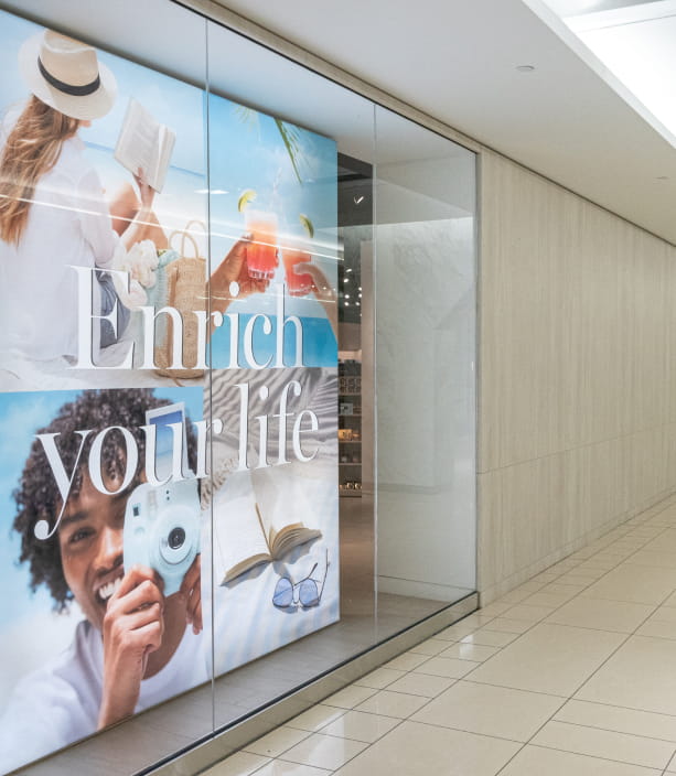 "A store window in a bright mall hallway displays a collage of lifestyle images including reading, cocktails, and photography, with the prominent text ""Enrich your life."""