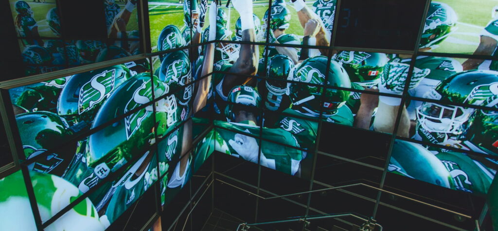 "An array of screens displays Canadian football players from the Saskatchewan Roughriders in green and white uniforms, some huddled together."