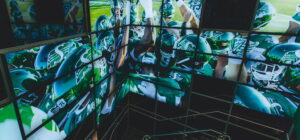 "An array of screens displays Canadian football players from the Saskatchewan Roughriders in green and white uniforms, some huddled together."