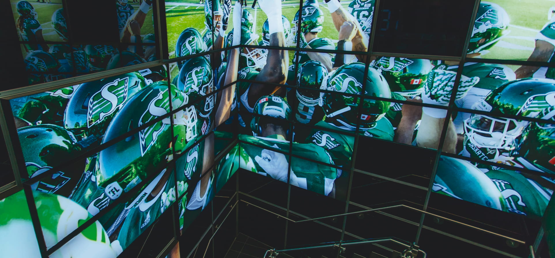 "An array of screens displays Canadian football players from the Saskatchewan Roughriders in green and white uniforms, some huddled together."