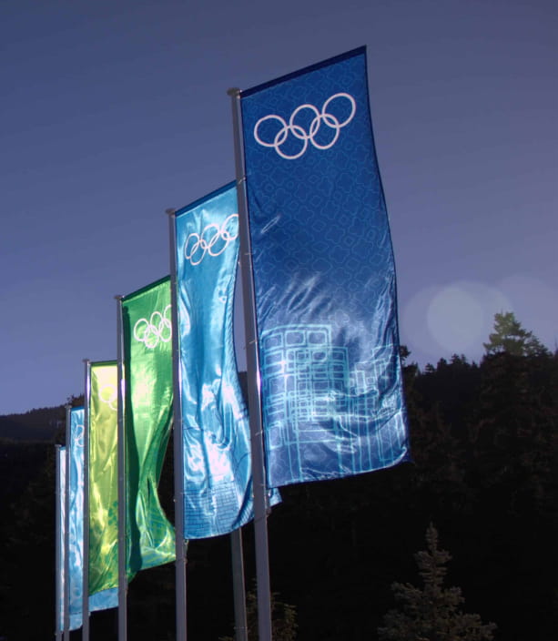 A row of blue and green Olympic flags with the Olympic rings logo fly against a clear blue sky and dark forest.