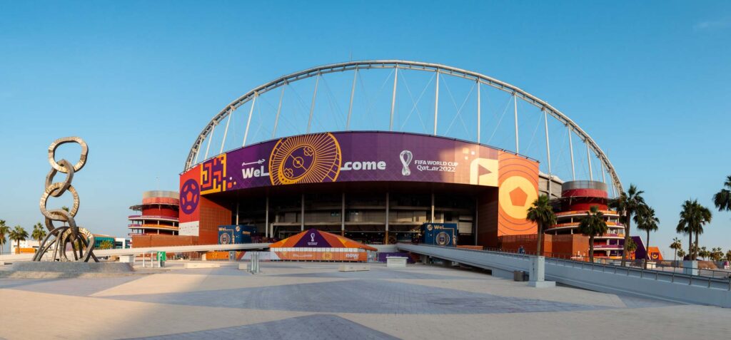 A large stadium exterior is decorated with ""Welcome"" and FIFA World Cup Qatar 2022 branding, with a prominent ring sculpture and palm trees in the foreground.