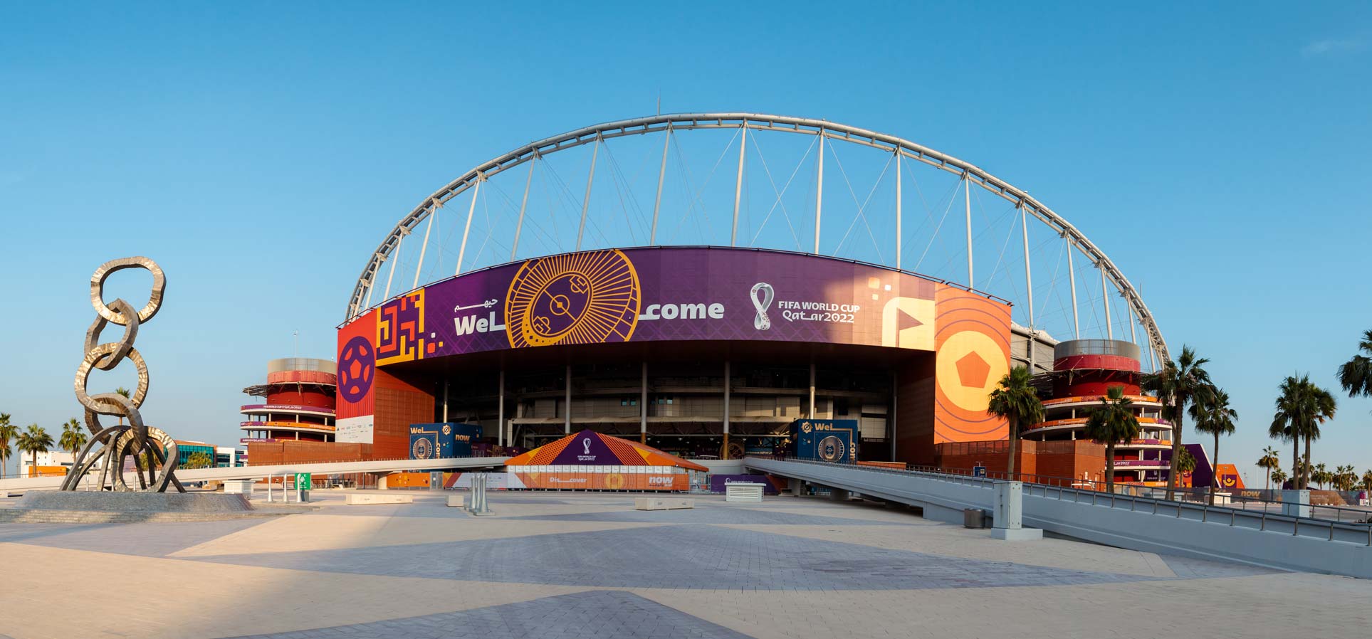 A large stadium exterior is decorated with ""Welcome"" and FIFA World Cup Qatar 2022 branding, with a prominent ring sculpture and palm trees in the foreground.