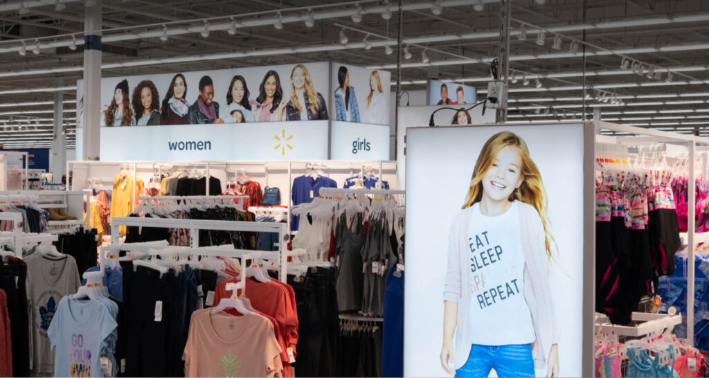 A brightly lit Walmart clothing department filled with racks of women's and girls' apparel under overhead signs featuring models.