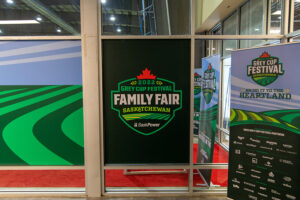 "A large green banner advertising the ""2022 Grey Cup Festival Family Fair Saskatchewan"" is displayed alongside other event signage and a field graphic."
