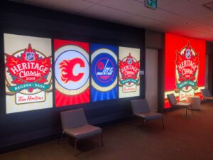 "Illuminated wall displays showcase the NHL Heritage Classic 2019, Calgary Flames, and Winnipeg Jets logos above lounge chairs in a hallway."
