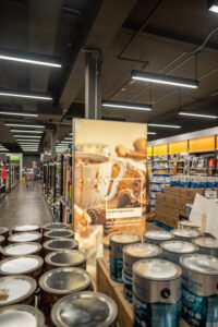 "Paint cans are displayed in the foreground of a brightly lit retail store, with a large sign advertising ""Double Fudge Brownie"" paint featuring dessert imagery."