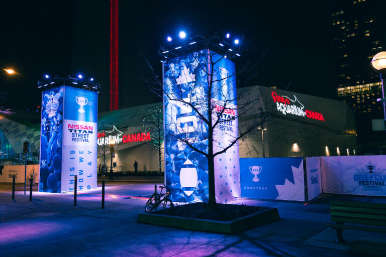 Brightly lit blue and white banners for the Nissan Titan Street Festival and Grey Cup Festival stand at night in front of Ripley's Aquarium of Canada and a red tower.