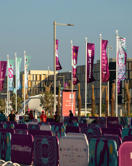 "A bustling outdoor area decorated with purple and turquoise FIFA World Cup Qatar 2022 flags and barriers, set against modern buildings."