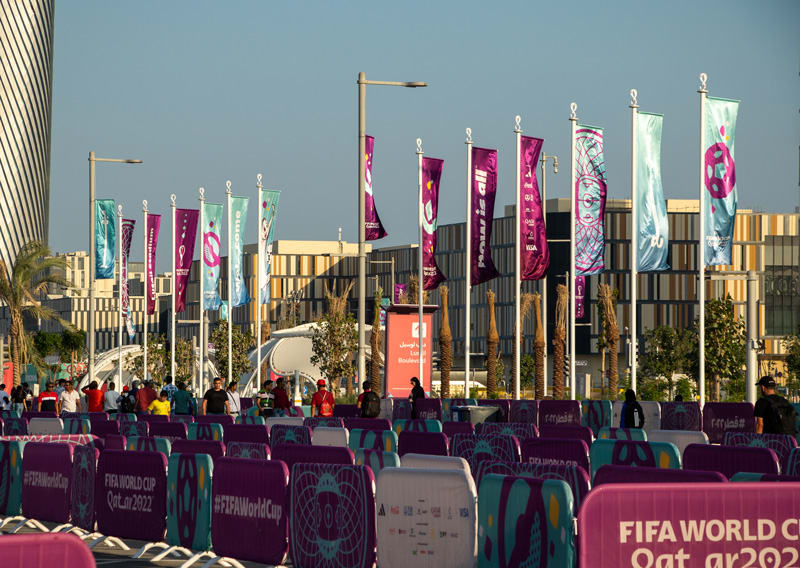 Numerous purple and teal flags and barriers branded for the FIFA World Cup Qatar 2022 line an outdoor urban area with people and modern buildings.