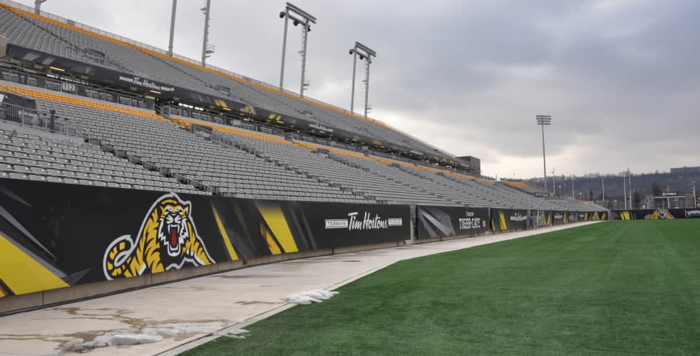 An empty outdoor sports stadium features rows of grey and yellow seating, a green turf field, and sponsor banners including the Hamilton Tiger-Cats logo.