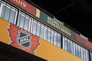 "A banner featuring the NHL logo on a red autumn leaf is displayed, with a ""Regina Saskatchewan 2019"" sign visible behind a railing."