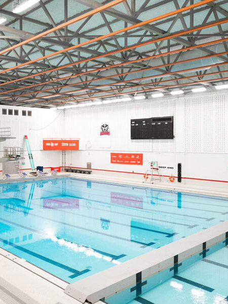 A bright, empty indoor swimming pool facility features diving platforms, marked lanes, and banners for the Canada Games.