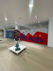 A mannequin in a vibrant patterned dress and teal boots stands on a white platform before a long wall mural featuring Matisse-inspired blue cutout figures and patterns on red and white backgrounds.