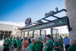 "A crowd of people in green shirts gathers at Gate 1 of a stadium entrance, featuring ""Proud Founding Partner"" signs, under a clear blue sky."