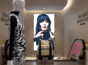 "A department store escalator descends past a large screen displaying a woman applying lipstick, with a mannequin visible in the blurred foreground."