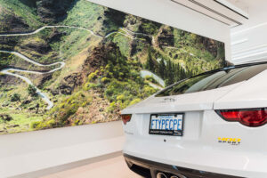 "A white Jaguar F-Type coupe with an ""FTYPCPE"" license plate is parked in front of a large wall mural depicting a winding mountain road."
