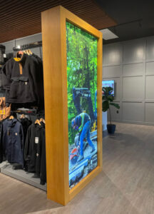 "A retail space showing clothing racks with workwear adjacent to a tall, wooden-framed digital display featuring a worker cutting concrete."