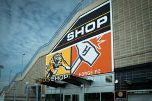 "Exterior of a building with a large ""SHOP"" sign above logos for the Tiger-Cats (tiger) and Forge FC (hammer)."