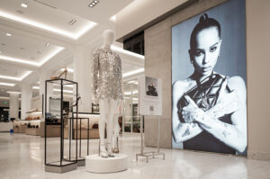 A brightly lit luxury department store features a mannequin in a silver sequined dress and a large black-and-white print of Zoë Kravitz holding a YSL bag.