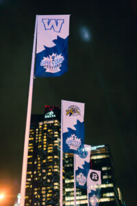 Nighttime view of multiple blue and white Grey Cup Festival flags with CFL team logos flying in front of illuminated city buildings.