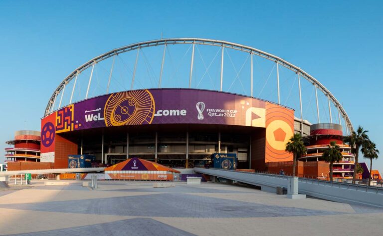 A large stadium featuring FIFA World Cup Qatar 2022 branding and a prominent arch structure under a clear blue sky.