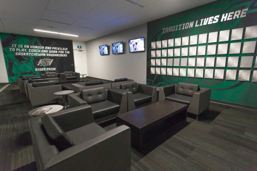 A lounge with dark seating, multiple TVs, and green Saskatchewan Roughriders branded walls featuring team mottos and framed achievements.