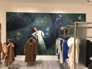 "A retail store with a large wall mural depicting two people in a dark, overgrown landscape, with racks of coats and other garments in the foreground."
