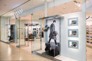 The modern glass storefront of an ECCO shoe store features shoes and bags displayed in its windows.