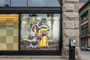 "A Louis Vuitton store window displays a vibrant fashion advertisement featuring two models in colorful clothing, alongside the brand logo."