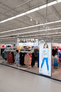 "Interior of a brightly lit clothing store with racks of women's and girls' apparel, featuring a large illuminated sign of a girl wearing an ""Eat Sleep Repeat"" shirt."