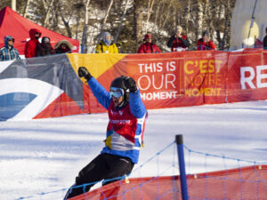 A snowboarder in a blue and black suit celebrates triumphantly on a snowy slope at the Red Deer 2019 games.