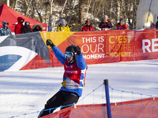 A snowboarder in a blue and black suit celebrates triumphantly on a snowy slope at the Red Deer 2019 games.