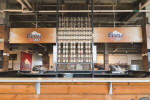 An empty Coors Banquet Bar featuring a long wooden counter and a tall display rack filled with glass mugs.