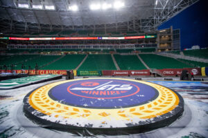 "A wide view of an empty outdoor stadium at night, with snow falling, highlighting a large Winnipeg Jets logo on a central stage and ""Heritage Classic 2019"" event banners."