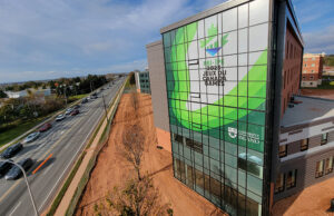 "A modern glass building with a large green banner for the ""PEI-IPE 2023 Jeux du Canada Games"" and the University of Prince Edward Island overlooks a multi-lane road and bare ground."