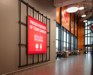 "A large illuminated red sign stating ""Proud Partner of Team Canada"" dominates a bright, spacious retail interior featuring tall windows, a maple leaf logo, and a display of barbecue grills."