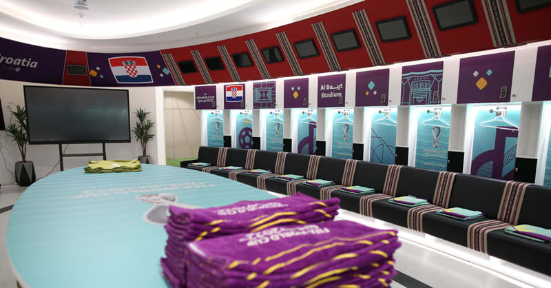 A FIFA World Cup Qatar 2022-themed room with a large table displaying purple merchandise, curved stadium-themed displays above black seating, and a large TV screen.