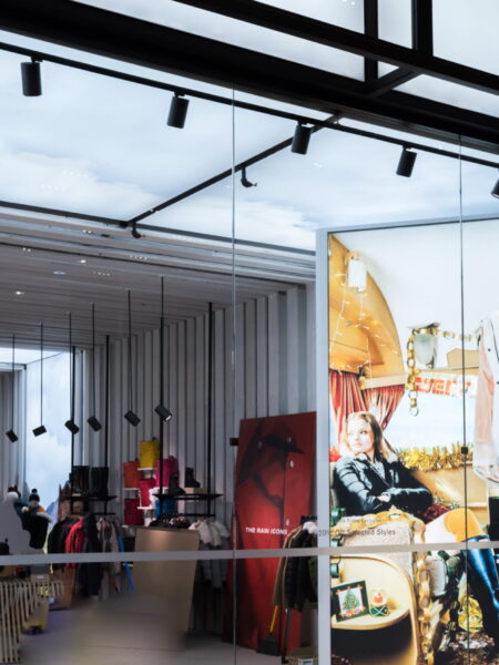 View into a modern fashion retail store through a glass facade, showcasing mannequins, clothing racks, and large promotional displays with models.