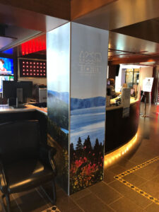A pillar featuring the G7 2018 Charlevoix logo and scenic landscapes stands in a modern lobby next to a reception desk.
