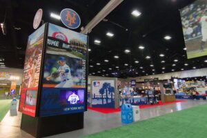 "An indoor MLB fan festival hall featuring large video displays of baseball players and stadiums, surrounded by various booths and a green turf floor."
