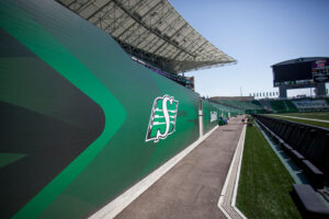 "Sideline view of a stadium with green Saskatchewan Roughriders branding, empty seats, and a scoreboard under a clear sky."
